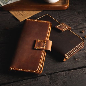 May include: Two leather wallets, one brown and one black, with contrasting stitching. The wallets are closed and lying on a dark wooden surface.