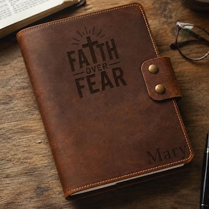 May include: A brown leather journal featuring a snap closure with two brass buttons. The cover is engraved with a cross icon and the text "FAITH OVER FEAR" above the name "Mary" in the bottom right corner, with visible stitching along the edges.