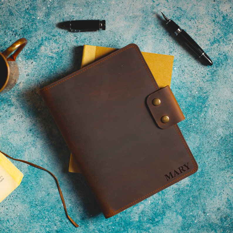 May include: A brown leather journal with a snap closure, personalized with the name "MARY" in a corner. The journal rests on a yellow notepad, with a pen and ink bottle nearby. The background is a textured teal surface.