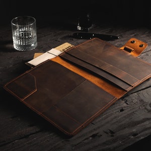 May include: Open, brown leather portfolio with multiple interior pockets and a snap closure. The portfolio has a lighter brown interior and is displayed open on a dark wooden surface. A glass of water and a pen are in the background.