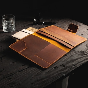 May include: Open brown leather folio with a yellow interior, featuring multiple pockets and a snap closure. The folio is placed on a dark wooden surface, with a glass and pen in the background. The folio is designed for documents and stationery.