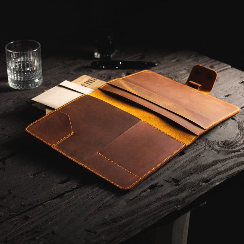 May include: Open brown leather folio with a yellow interior, featuring multiple pockets and a snap closure. The folio is placed on a dark wooden surface, with a glass and pen in the background. The folio is designed for documents and stationery.