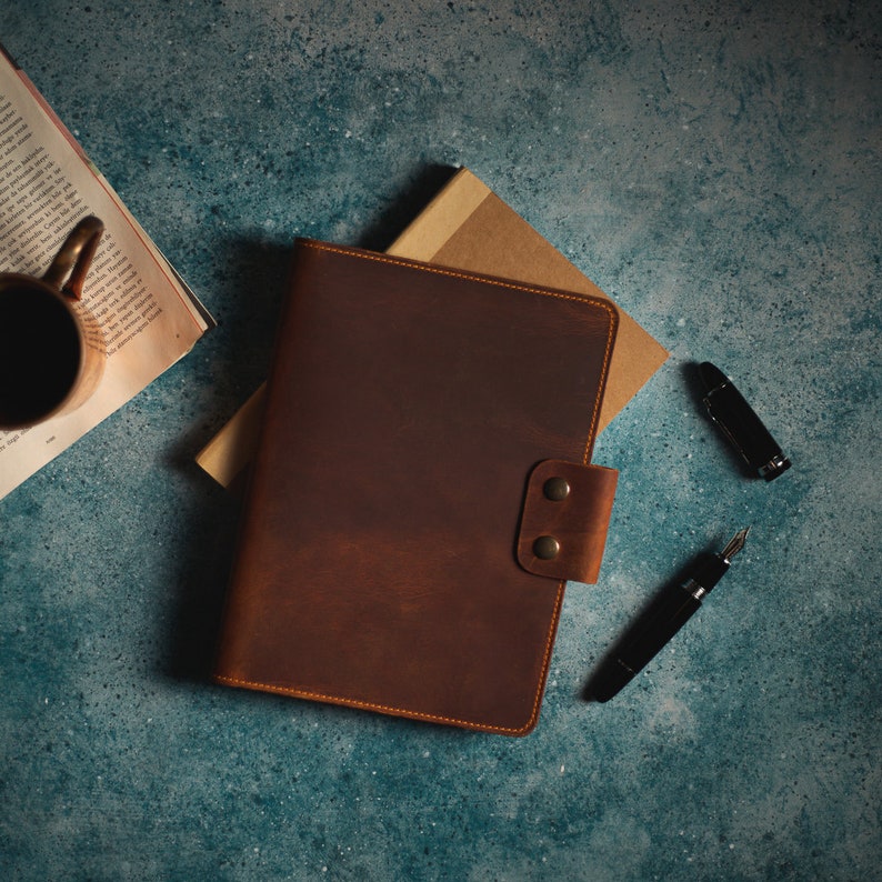 May include: A brown leather journal with a button clasp, a black fountain pen, and a black pen are arranged on a textured blue surface. A cup of coffee and a book are also visible, suggesting a workspace or study setting.