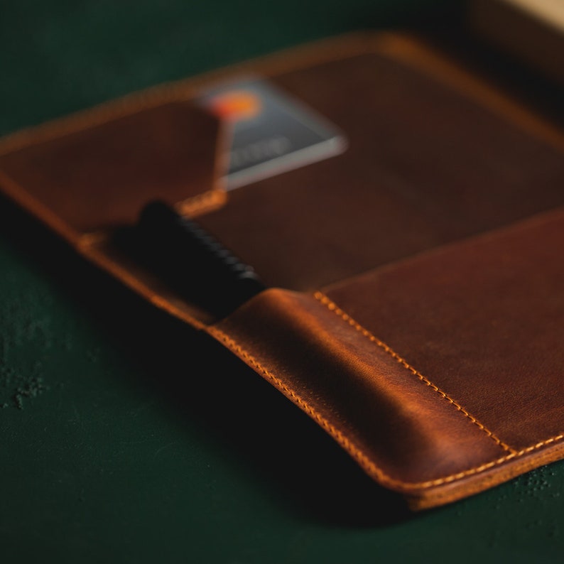 May include: Open brown leather wallet with a pen in a side pocket and a credit card in a slot. The wallet has a stitched border and is set against a dark green background.