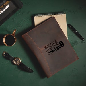 Pu&ograve; includere: Un portafoglio in pelle marrone con un bordo cucito e il testo "YOUR LOGO" in rilievo sul davanti. Il portafoglio &egrave; appoggiato su una superficie verde con un taccuino, una penna e un orologio.