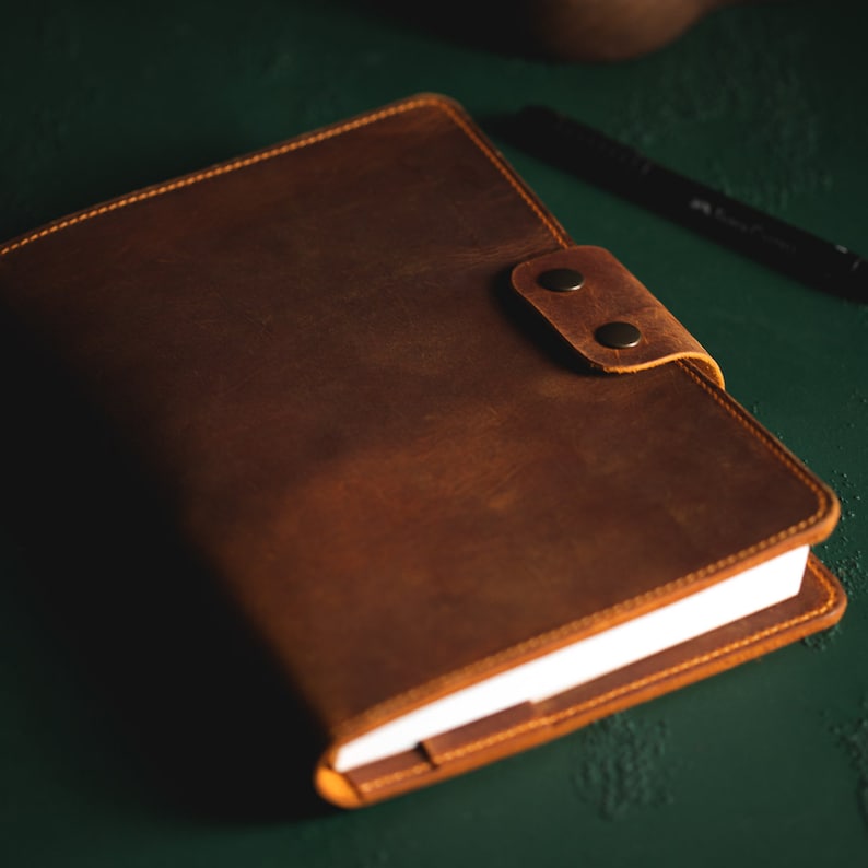 May include: A brown leather journal with a button closure. The journal has a textured surface and visible stitching around the edges. The pages are white, and a black pen rests nearby. The journal is on a dark green surface.