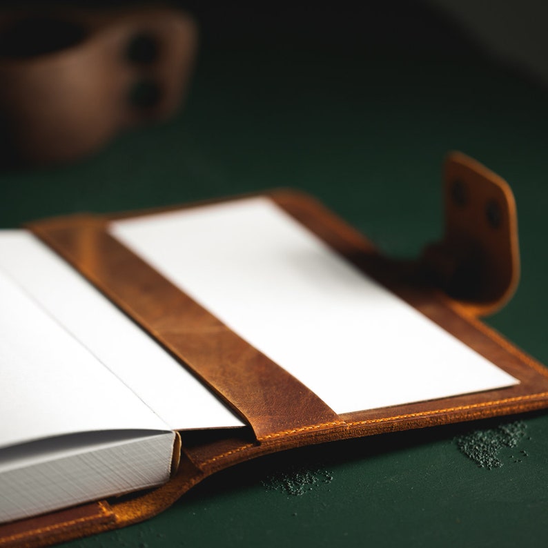 May include: Open brown leather journal with white pages. The journal is on a dark green surface. The leather has a distressed look, with visible stitching along the edges. A brown leather strap secures the journal.