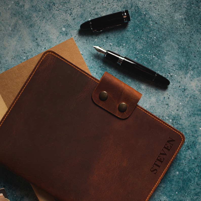 May include: A brown leather journal with the name "STEVEN" embossed on the cover. The journal is closed with a flap and two brass-colored snaps. A black fountain pen and a pen cap are placed on a blue-green surface.