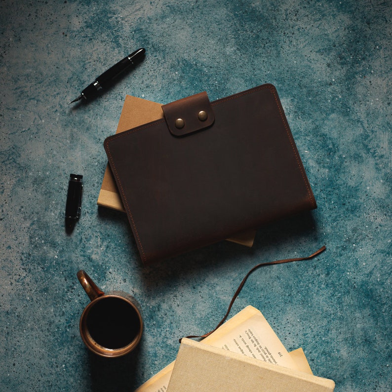 May include: A dark brown leather portfolio with a button clasp, resting on a stack of books. A black fountain pen and a coffee mug are also visible. The items are arranged on a textured teal surface.