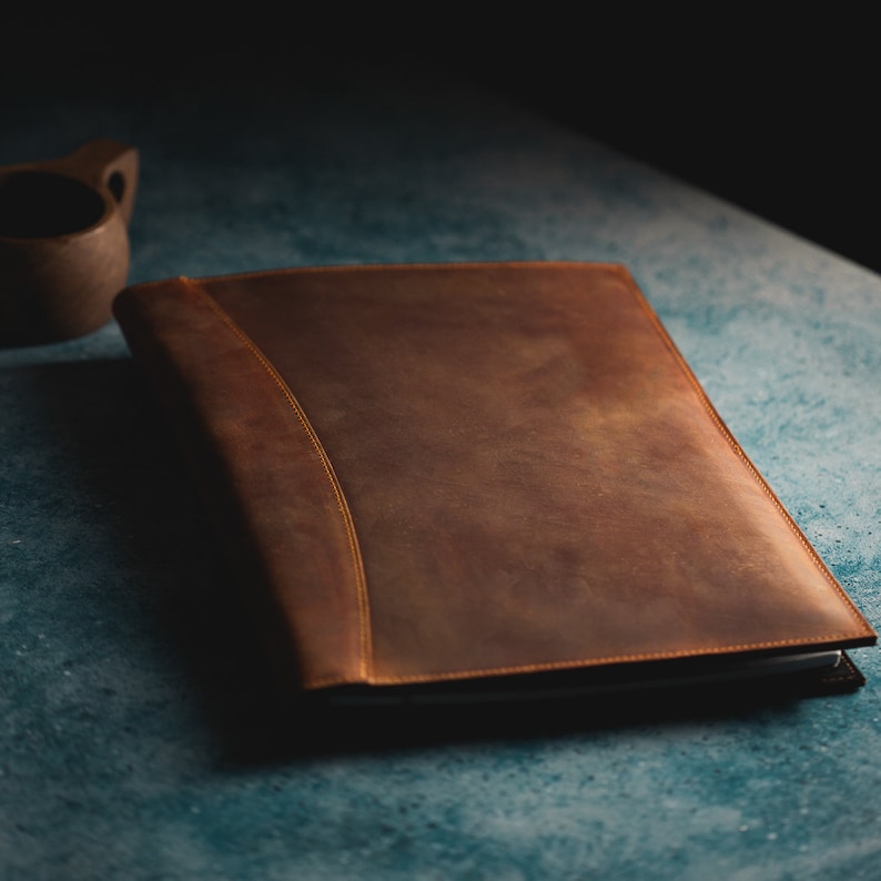 Pu&ograve; includere: Una cartella in pelle marrone con un bordo cucito e un design curvo sottile. La cartella &egrave; chiusa e giace piatta su una superficie blu.