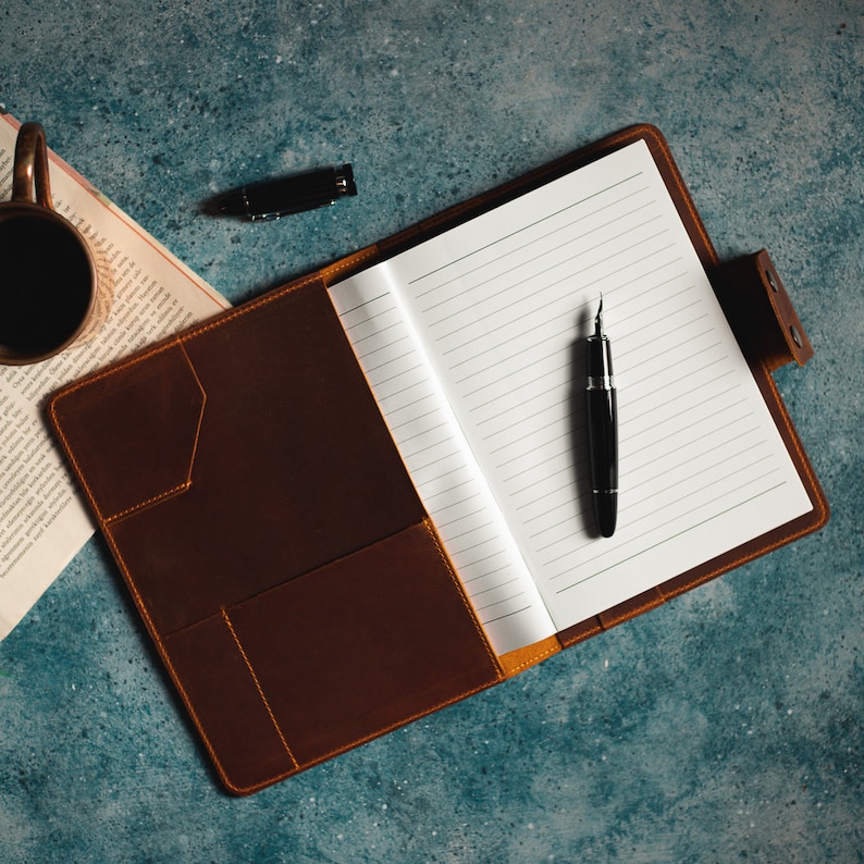 May include: Open brown leather journal with lined pages and a black fountain pen. The journal is on a blue surface, with a cup of coffee and a pen cap visible in the background. The journal has pockets.
