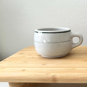 May include: Two white ceramic mugs with a black stripe near the rim and a thin black line around the middle. The mugs have a rounded shape and small handles. They are on a light-coloured wooden surface.