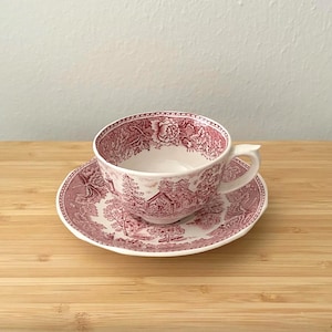 May include: A white teacup and saucer set with a red floral and landscape pattern. The teacup has a handle and sits on a matching saucer. The set is on a wooden surface.