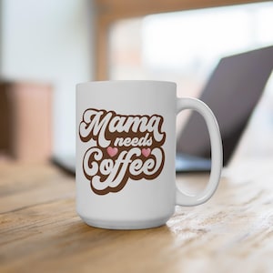 May include: White ceramic coffee mug with the text "Mama needs Coffee" in brown script. Two pink hearts are between the words "Coffee" and "needs". The mug has a large handle and is on a wooden surface.