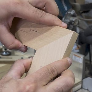 May include: Close-up of two pieces of light-colored wood being joined at a right angle. The wood has been cut at an angle to fit together. A miter saw and other woodworking tools are in the background.