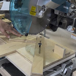 May include: A close-up shot of a miter saw with a circular blade cutting through light-colored wood. The saw has a clear safety guard and is set up for precise angled cuts. A hand is guiding the wood.