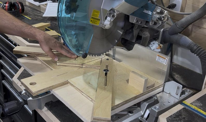 May include: A close-up shot of a miter saw with a circular blade cutting through light-colored wood. The saw has a clear safety guard and is set up for precise angled cuts. A hand is guiding the wood.