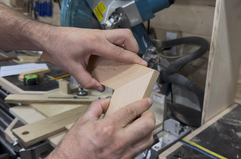 May include: Close-up of two pieces of light-colored wood being joined at a right angle. The wood has been cut at an angle to fit together. A miter saw and other woodworking tools are in the background.