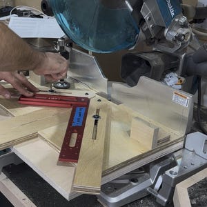 May include: A miter saw with a blue blade guard and a red angle measuring tool. Several pieces of light-colored wood are positioned on the saw's platform, ready for cutting. The scene suggests woodworking or carpentry.