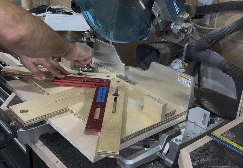 May include: A miter saw with a blue blade guard and a red angle measuring tool. Several pieces of light-colored wood are positioned on the saw's platform, ready for cutting. The scene suggests woodworking or carpentry.