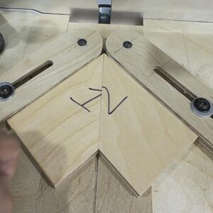 May include: A woodworking jig with two pivoting wooden arms, each with a metal bolt and washer. A piece of light-colored wood is positioned between the arms, with the handwritten letters "H2" in black marker. The jig is on a light wood surface.
