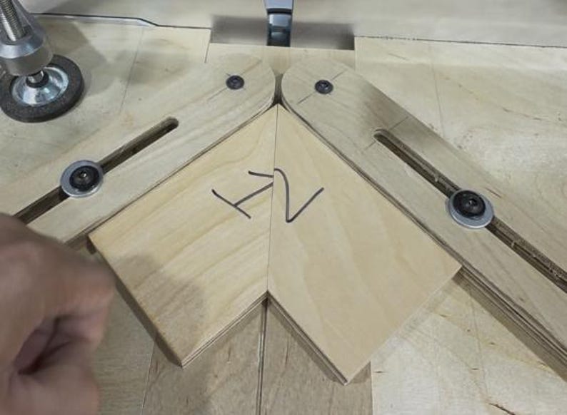 May include: A woodworking jig with two pivoting wooden arms, each with a metal bolt and washer. A piece of light-colored wood is positioned between the arms, with the handwritten letters "H2" in black marker. The jig is on a light wood surface.