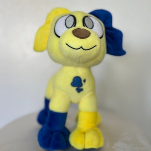 May include: A yellow plush dog toy with blue accents. The dog has large white eyes, a brown nose, and a blue bow on its ear. The dog is standing on a white surface.
