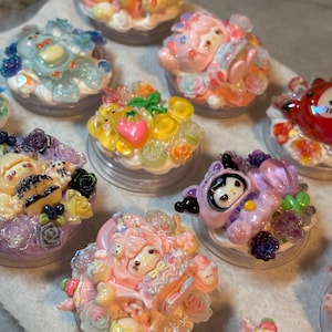 May include: A collection of circular phone grips decorated with various colorful resin cartoon characters, miniature flowers, and translucent fruit shapes. Each grip features a unique arrangement of glossy, multi-colored embellishments.
