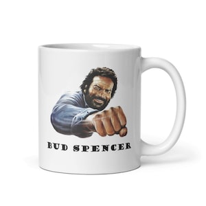 Può includere: Tazza in ceramica bianca con un'immagine di Bud Spencer, un famoso attore italiano, con barba e baffi, che indossa una camicia blu e sferra un pugno con il suo pugno destro. Il testo "BUD SPENCER" è stampato in nero sotto l'immagine.