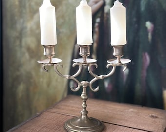 May include: A brass candelabra with three candle holders and a decorative scrolled design. The candelabra is holding three white candles.
