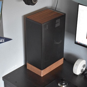 Puede incluir: Una caja de ordenador negra con la parte superior e inferior de madera está sobre un escritorio. La caja tiene forma rectangular con un diseño moderno. Un monitor de ordenador y otros accesorios tecnológicos son visibles en el fondo.