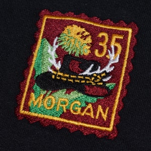Op de afbeelding: Een geborduurde patch in rood en goud met een groene boom, geweien en een hoed. De patch heeft de tekst "35 MORGAN" in gouden letters.