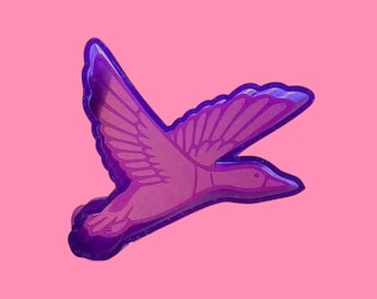 May include: A purple and pink duck pin in flight, set against a pink background. The pin features a detailed outline and wing design, with a glossy finish. The duck is a stylized representation.