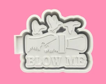 May include: A white plastic cookie cutter in the shape of a duck call with the words "BLOW ME" in bold letters. The design includes ducks in flight and a silhouette of a person hunting.
