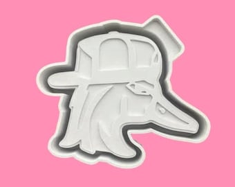 May include: A white plastic cookie cutter in the shape of a duck wearing a baseball cap. The duck's head is turned to the side, and the cap is detailed with a curved brim. The cutter is set against a pink background.