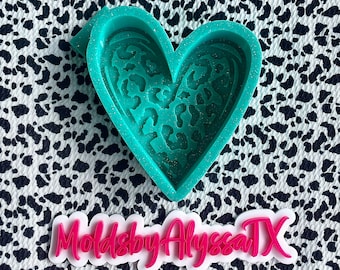 May include: A teal blue heart-shaped silicone mold with a leopard print design. The mold is on a black and white patterned surface. The text "MoldsbyAlyssaTX" is visible in the lower right corner of the image.