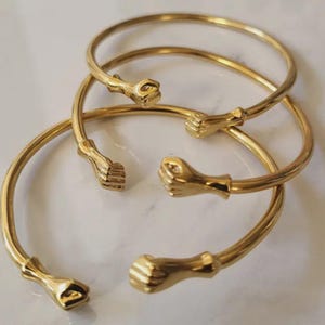 May include: Gold-colored metal bangles with a fist design. The open-ended bracelets feature a smooth, polished finish and a fist at each end. The bangles are displayed on a white surface.