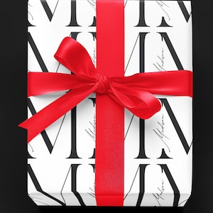 May include: A white gift box with a red ribbon tied in a bow. The box is wrapped in paper with a black and white pattern of the letter "M" and the word "Madison".