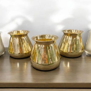 May include: Three shiny, gold-coloured metal pots with a rounded base and a wide, flared rim. The pots have horizontal etched lines around the body. They are arranged on a wooden surface against a white background.