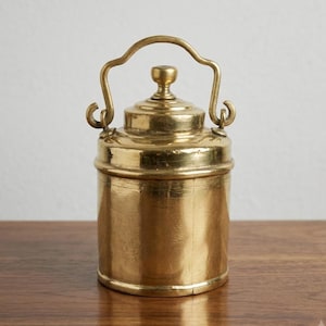 May include: A cylindrical brass container with a polished metallic finish, featuring a fitted lid with a central knob and a curved, hinged handle attached to two side loops. The vessel has a smooth, solid construction with a warm golden tone.