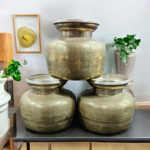 May include: Three antique brass pots with wide bodies and flared rims are stacked. The pots have a tarnished, aged appearance with horizontal etched lines around the body. The pots are displayed on a dark surface, with plants and artwork in the background.