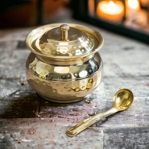 Puede incluir: Un recipiente de metal dorado martillado con tapa y una cuchara pequeña. El recipiente tiene la palabra "food" grabada en el lateral. La cuchara y el recipiente están sobre una superficie gris y desgastada.