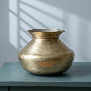 May include: A brass pot with a hammered texture, featuring a rounded body and a flared rim. The pot has a golden hue and sits on a teal surface. The background is a white wall with shadows.