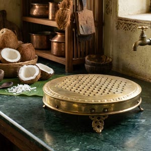 May include: A round, brass kitchen grater with small holes and decorative feet sits on a green countertop. The grater is in front of a basket of coconuts, coconut halves, and a wooden shelf with copper pots.