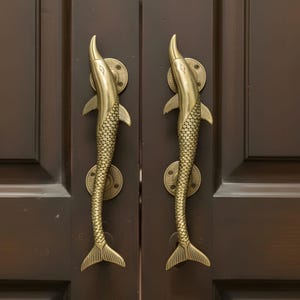 May include: A pair of brass dolphin-shaped door handles fixed to a dark brown wooden door. The handles feature scale and fin detailing, and are mounted with circular plates. The door handles are approximately 30 cm in length.