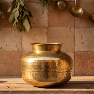 May include: A hammered brass pot with a wide, rounded body and a flared rim. The pot has a textured surface and reflects the warm light of the room. The pot is sitting on a wooden surface.