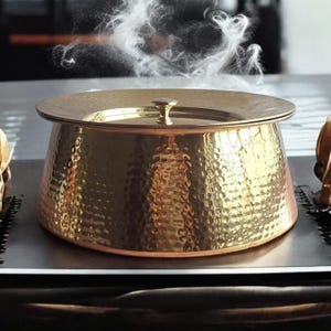 May include: A gold hammered metal pot with a lid. The pot is sitting on a black surface and steam is rising from it.