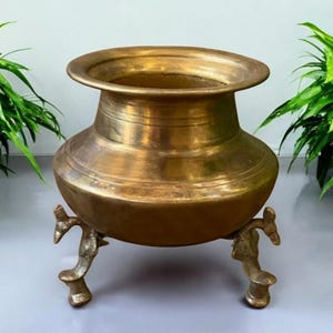Peut inclure: Un pot en laiton vintage avec un large bord et un corps arrondi, soutenu par trois pieds ornés. Le pot a une teinte dorée chaude et est probablement utilisé pour la cuisine ou la décoration. Les dimensions du pot ne sont pas visibles.