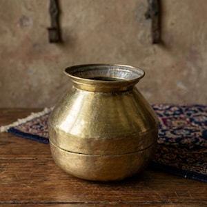 Puede incluir: Una olla de latón vintage con un cuerpo ancho y redondeado y un borde acampanado. La olla tiene un tono dorado cálido y una apariencia ligeramente envejecida. Se encuentra sobre una superficie de madera, con una alfombra estampada visible en el fondo. El diseño de la olla sugiere que puede usarse con fines decorativos.