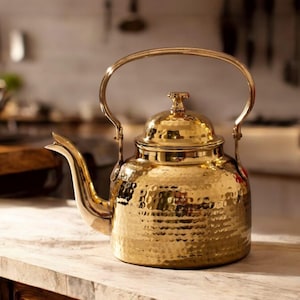 May include: A gold hammered metal kettle with a lid and a curved handle. The kettle has a textured surface and a spout.
